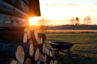 Stacked firewood logs and wheelbarrow silhouetted against golden sunset in countryside, warm evening light illuminating rural farm scene with dramatic lens flare.
