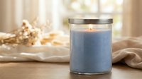 Blue scented candle burning with warm flame in glass jar on wooden surface with soft natural lighting creating cozy home atmosphere for relaxation.