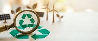 Magnifying glass revealing green recycling symbol with wind turbines and solar panel in blurred background, representing environmental sustainability and renewable energy solutions.