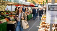 Young woman shopping at outdoor farmers market with fresh produce and organic vegetables, holding reusable bag and smartphone for sustainable lifestyle.