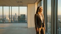 Professional businesswoman in suit standing by office windows overlooking city skyline at sunset, corporate success and leadership concept.