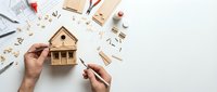 Hands assembling wooden house model kit with construction tools and blueprints on white background for DIY home building project.