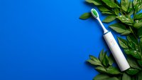 Electric toothbrush with green fresh leaves on bright blue background, minimalist dental hygiene composition for healthy lifestyle concept.