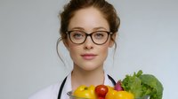 Young female healthcare professional in white coat and glasses holding fresh vegetables and fruits, promoting healthy eating and nutrition guidance.