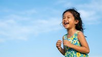 Young Asian girl with dark hair wearing a colorful floral sleeveless dress, laughing joyfully outdoors against a bright blue sky with white clouds, expressing happiness and childhood joy.