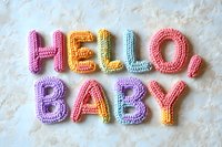 Colorful crocheted letters spelling HELLO BABY in rainbow colors on white textured background for nursery decor and baby shower celebrations.