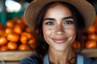 Young Hispanic woman farmer wearing straw hat and denim overalls smiling at farmers market with fresh oranges in background, close up portrait.