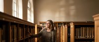 Young woman browsing books in vintage library with wooden shelves and warm natural lighting from tall windows creating peaceful academic atmosphere.