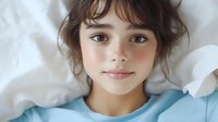 Young girl with freckles and dark hair lying on white pillows, wearing light blue pajamas, smiling gently at camera with natural morning light.