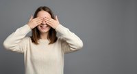Young woman covering eyes with hands wearing beige sweater on gray background expressing surprise shame or playing peek-a-boo game in studio portrait.