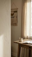 Cozy reading corner with wooden table, coffee mug, open book and macrame wall hanging in warm natural light creating peaceful home atmosphere.