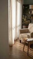 Cozy reading corner with beige armchair, wooden coffee table, book and mug in warm sunlit living room with natural light streaming through curtains.