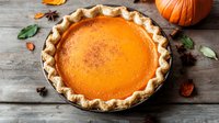 Fresh homemade pumpkin pie with flaky crust and smooth orange filling on rustic wooden background with autumn decorative elements and spices.