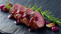 Seared duck breast sliced medium rare with cranberry sauce, fresh rosemary garnish, and roasted garlic on dark slate background, elegant restaurant presentation.
