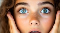Young woman with striking blue eyes and natural freckles touching face with hands showing surprise expression for beauty skincare cosmetics advertising.
