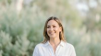 Young Caucasian woman with shoulder-length blonde hair smiling at camera wearing white button-up shirt against blurred green foliage background for portrait and professional use.