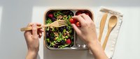 Overhead view of hands eating fresh salad with vegetables and greens from a container using wooden utensils on white surface.