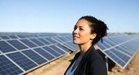 Young woman of mixed ethnicity wearing dark business attire standing in solar panel farm, looking toward horizon. Professional in renewable energy sector surveying sustainable power installation.