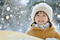 Cheerful Asian child wearing knit winter hat and yellow coat smiling outdoors during snowfall, capturing pure joy and seasonal magic against blurred bokeh background.