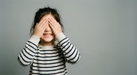 Young Asian girl covering eyes with hands wearing striped shirt against gray background expressing surprise shame or playing peek-a-boo game.