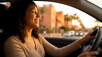 Woman with long dark curly hair, Hispanic ethnicity, driving car during golden hour sunset with urban buildings and palm trees blurred in background. Suitable for automotive, transportation, and lifestyle content.