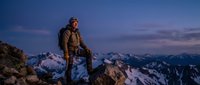 Male mountaineer with backpack standing on rocky summit during twilight with snow-capped mountain peaks in background. Adventure hiking concept.