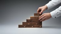 Person with light skin tone wearing white business shirt placing wooden blocks with checkmarks in pyramid formation, representing success, growth, and achievement in corporate strategy and planning.