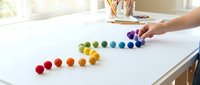 Child arranging colorful rainbow felt balls in row on white table for early learning color sorting educational activity at home.