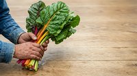 Close-up of male hands holding fresh rainbow chard with vibrant green leaves and colorful stems on wooden surface for cooking and healthy eating.