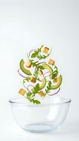 Fresh salad ingredients falling into glass bowl with avocado slices, arugula leaves, red onion rings and croutons on white background.