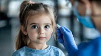 Young caucasian girl with pigtails receiving medical ear examination from pediatrician wearing protective blue gloves and face mask during routine checkup at clinic.