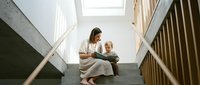 Young mother with toddler son sitting on staircase reading book together in bright modern home interior for family bonding and early childhood education.