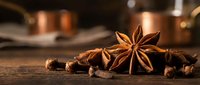 Star anise and cloves scattered on rustic wooden surface with warm copper cookware in blurred background creating cozy kitchen atmosphere for culinary designs.