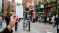 Hand holding blank white card in busy urban street with blurred pedestrians and storefronts in background, suitable for advertising and promotional campaigns.