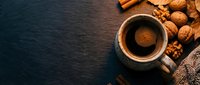 Overhead view of steaming coffee in ceramic mug surrounded by walnuts, almonds, and knitted fabric on dark slate surface for beverage and food styling.