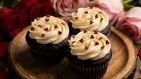 Chocolate cupcakes with cream frosting and dried cranberries on wooden board, surrounded by pink roses for romantic celebration or Valentine's Day dessert.