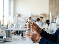 Hand holding clear glass bottle with liquid in laboratory setting with blurred scientists working in background, suitable for research, chemistry, pharmaceutical, and scientific education content.