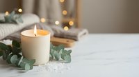 Burning white candle with eucalyptus leaves on marble surface creating cozy hygge atmosphere with warm bokeh lights in background for relaxation.