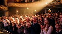 Enthusiastic audience applauding in elegant theater with ornate architecture and warm lighting during live performance or cultural event.