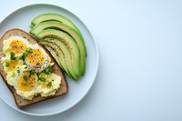 Sliced avocado and soft boiled egg on whole grain toast garnished with fresh herbs and black pepper, served on white ceramic plate against light background.