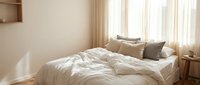 Cozy minimalist bedroom with white linen bedding, neutral pillows, sheer curtains, and warm natural light creating a serene, relaxing atmosphere.