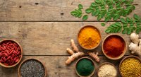 Colorful spices and herbs arranged in wooden bowls on rustic table with fresh moringa leaves and turmeric roots for cooking and seasoning.