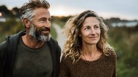 Mature Caucasian couple with gray-haired bearded man and blonde woman in sweaters smiling outdoors at sunset in natural setting.