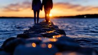 Silhouette of couple standing on pier at sunset, with sun rays breaking through between them over tranquil water.