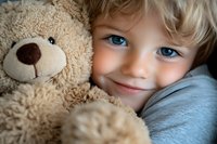 Smiling young Caucasian child with bright blue eyes hugging soft beige teddy bear, wearing light blue sweater, close-up portrait showing innocence and joy.