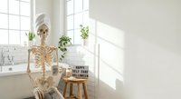 Human skeleton model sitting in bright modern bathroom with white subway tiles and natural light for medical education and anatomy study.