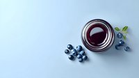 Fresh blueberry jam in glass jar with scattered berries and mint leaves on light blue background, perfect for food packaging design.
