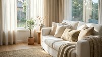 Cozy modern living room interior with beige sofa, decorative pillows, knitted throw blanket, potted plant and natural sunlight through large windows.