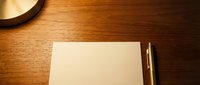 Blank notepad with pen on wooden desk surface next to desk lamp, ready for writing, planning, or business correspondence.