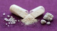 Open capsules spilling white powder supplement on purple textured surface with granules scattered around for health and wellness concepts.